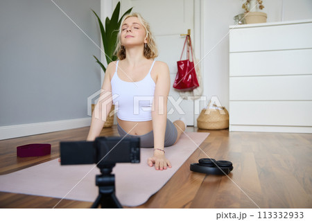 Portrait of young blogger, yoga content creator, showing exercises, recording video of herself working out at home on rubber mat Portrait of young blogger, yoga content creator, showing exercises, recording video of herself working out at home on rubber mat 113332933