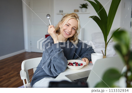 Portrait of smiling blond young woman, eating in front of laptop, watching videos online while having breakfast, enjoying dessert, sitting in bedroom Portrait of smiling blond young woman, eating in front of laptop, watching videos online while having breakfast, enjoying dessert, sitting in bedroom 113332934