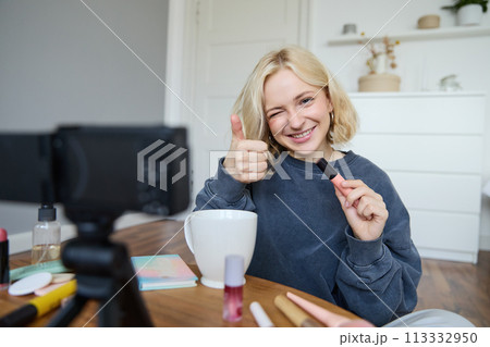 Portrait of smiling, beautiful young woman, lifestyle blogger recording a video in her room, sitting in front of camera on stabiliser, showing thumbs up and wink face, recommending mascara Portrait of smiling, beautiful young woman, lifestyle blogger recording a video in her room, sitting in front of camera on stabiliser, showing thumbs up and wink face, recommending mascara 113332950