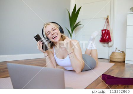 Portrait of woman lying on rubber yoga mat in room, wearing headphones, listens to music, using laptop and smartphone 113332969
