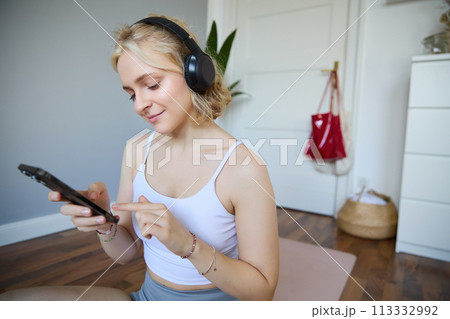Portrait of young sporty woman in wireless headphones, sitting with smartphone on rubber mat, using workout training app, fitness application on her phone, listening in earphones Portrait of young sporty woman in wireless headphones, sitting with smartphone on rubber mat, using workout training app, fitness application on her phone, listening in earphones 113332992
