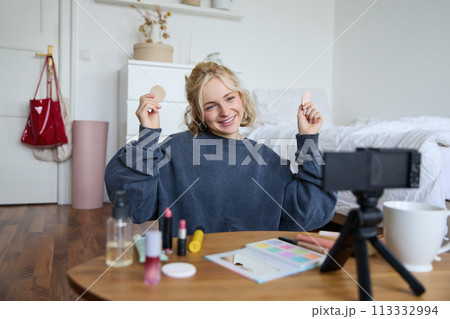 Portrait of beautiful and stylish young woman, vlogger recording video on her digital camera in a room, showing beauty products, making makeup tutorial for followers on social media 113332994