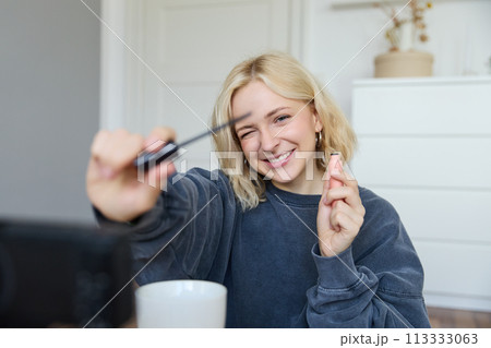 Portrait of young smiling woman in her room, recording video on camera, lifestyle vlog for social media, holding mascara, reviewing her makeup beauty products, showing how to use cosmetics 113333063