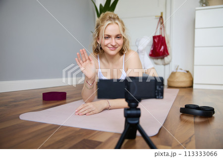 Portrait of smiling fitness instructor, social media content creator recording a vlog about sport, using rubber yoga mat, waving hand and looking at digital video camera 113333066