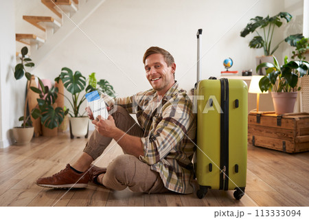 Portrait of male model with suitcase, sitting on floor, holding flight tickets and passport, going abroad, waiting for a taxi to airport Portrait of male model with suitcase, sitting on floor, holding flight tickets and passport, going abroad, waiting for a taxi to airport 113333094