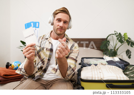 Happy tourist, young man shows passport and tickets, goes on holiday, sits with packed suitcase on bed, plans summer vacation Happy tourist, young man shows passport and tickets, goes on holiday, sits with packed suitcase on bed, plans summer vacation 113333105