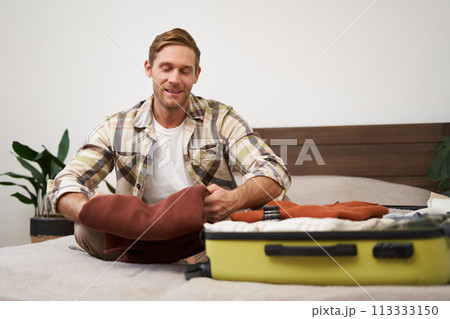 Portrait of handsome young man, tourist unpacking clothes in hotel room, staying in hostel on vacation, travelling, sitting with luggage on bed 113333150