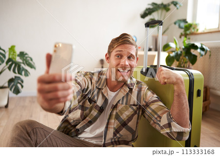 Funny handsome guy, tourist flexing biceps, goes on vacation, sits with suitcase, takes selfies on smartphone 113333168