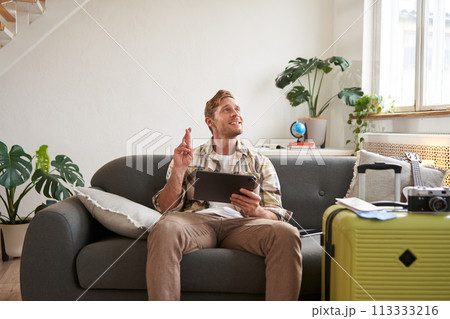 Excited young with tablet, sits in living room with packed suitcase, cross fingers for good luck, making wish, hoping for something Excited young with tablet, sits in living room with packed suitcase, cross fingers for good luck, making wish, hoping for something 113333216