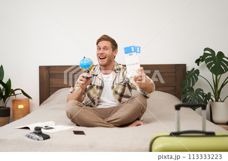 Image of happy smiling man, cheerful tourist sits with globe and plane tickets, goes on holiday, sits with suitcase, excited about his trip, going on holiday 113333223