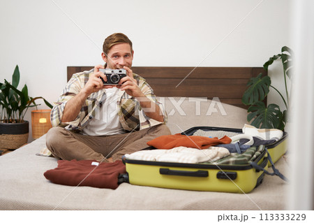 Portrait of tourist, man sits with suitcase on bed, holds camera, packs suitcase with clothes, goes on holiday, prepares for summer vacation, tourist travels abroad 113333229