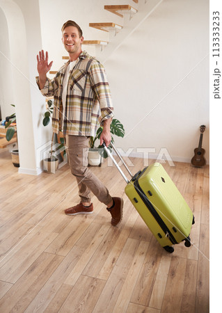 Vertical portrait of happy guy, tourist leaving the apartment with suitcase, waving hand at camera, saying hello or goodbye Vertical portrait of happy guy, tourist leaving the apartment with suitcase, waving hand at camera, saying hello or goodbye 113333233