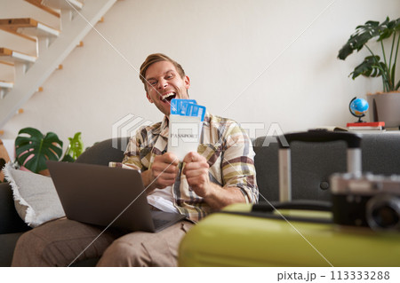 Portrait of happy handsome guy, shows his flight tickets, booked a trip for summer holiday, sits with laptop in living room, smiles at camera Portrait of happy handsome guy, shows his flight tickets, booked a trip for summer holiday, sits with laptop in living room, smiles at camera 113333288