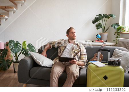 Image of young man with digital tablet, tourist sitting on sofa with suitcase packed and ukulele, thinking, looking aside thoughtful, planning holiday trip 113333292