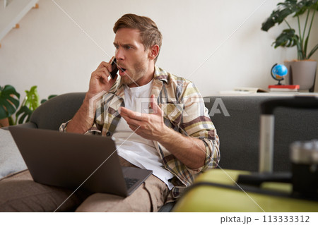 Portrait of angry man arguing over the phone, has laptop opened on his laptop, sits near suitcase, complaining at travel agency Portrait of angry man arguing over the phone, has laptop opened on his laptop, sits near suitcase, complaining at travel agency 113333312