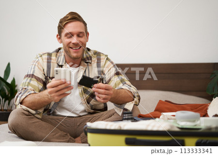 Smiling tourist, man with suitcase, sits on bed, pays online, books a hotel, holds credit card and mobile phone, uses an app to purchase in internet 113333341