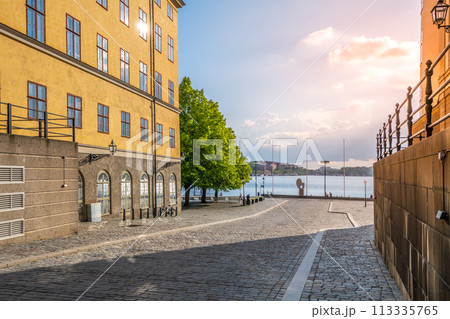 Warm sunlight bathes the historic buildings and cobblestone streets at Riddarholmen, overlooking the tranquil waterfront as day transitions to evening. Stockholm, Sweden 113335765