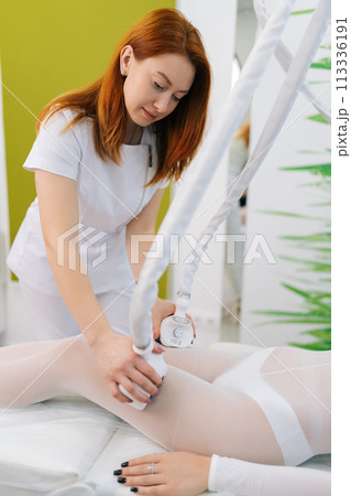 Vertical portrait of female doctor manipulating hardware cosmetology LPG gadget for lifting body in spa center. Female client wearing special white suit receiving anti-cellulite massage in salon. Vertical portrait of female doctor manipulating hardware cosmetology LPG gadget for lifting body in spa center. Female client wearing special white suit receiving anti-cellulite massage in salon. 113336191