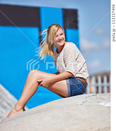 Happy, girl and teenager relax outdoors with feet in skatepark in summer with casual fashion or style. Person, sitting and smile on ramp, waiting or watching skating outdoor event on holiday 113337404