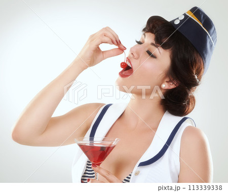 Cocktail, stewardess and woman eating cherry in studio isolated on white background. Red martini, glass and air hostess enjoy alcohol fruit, vintage pin up girl or flight attendant travel on journey Cocktail, stewardess and woman eating cherry in studio isolated on white background. Red martini, glass and air hostess enjoy alcohol fruit, vintage pin up girl or flight attendant travel on journey 113339338
