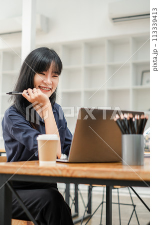 Cheerful young woman with pen in hand working on her laptop in a bright cafe, with a coffee cup on the table. 113339413