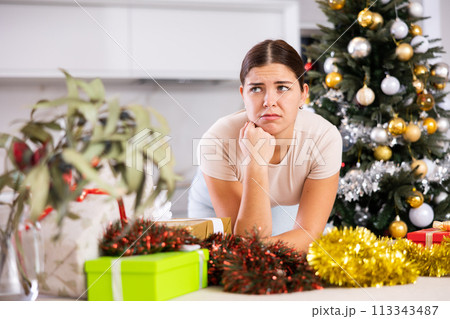 Emotional sad young female next to packed gift box with bow near and decorated Christmas tree at home Emotional sad young female next to packed gift box with bow near and decorated Christmas tree at home 113343487