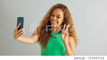Happy woman, peace sign and selfie with kiss for social media, vlog or memory on a gray studio background. Female person with smile on mobile smartphone for photography or picture on mockup space 113347122