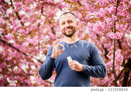 Man allergic suffering from seasonal allergy in blossoming garden at spring. Handsome smiling man holding allergy pill, making 'OK' hand sign in front of blooming tree. Spring allergy concept. Man allergic suffering from seasonal allergy in blossoming garden at spring. Handsome smiling man holding allergy pill, making 'OK' hand sign in front of blooming tree. Spring allergy concept. 113350650