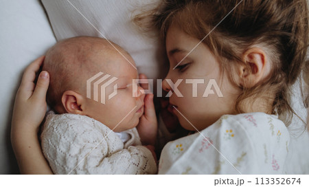 Portrait of big sister cuddling newborn, little baby. Girl lying with her new sibling in bed, closed eyes. Sisterly love, joy for new family member. Portrait of big sister cuddling newborn, little baby. Girl lying with her new sibling in bed, closed eyes. Sisterly love, joy for new family member. 113352674
