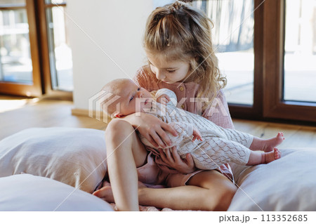 Portrait of big sister holding newborn sister. Girl carefully cuddling small baby. Sisterly love, joy for new family member. 113352685