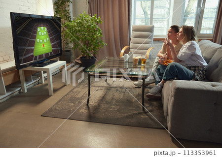 Tense game moment. Two girls, friends sitting in living room at home, eating snack and watching online football match on TV Tense game moment. Two girls, friends sitting in living room at home, eating snack and watching online football match on TV 113353961