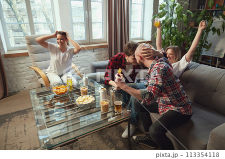 Scoring winning goal. Young people, friends sitting in living room at home, emotionally watching football match online translation. Scoring winning goal. Young people, friends sitting in living room at home, emotionally watching football match online translation. 113354118
