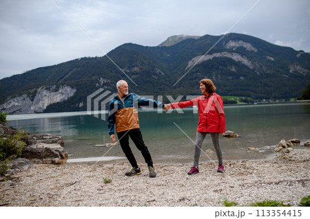Active elderly couple hiking together in spring mountains, walking on pebble beach of mountain lake. Senior tourists enjoying nature. 113354415