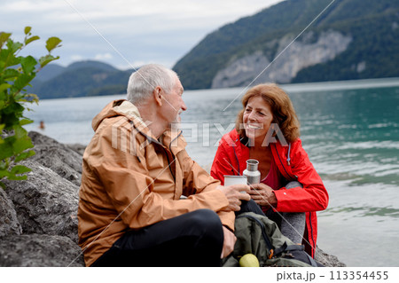 Active elderly couple hiking together in mountains. Drinking coffee and having healthy snack, to get energy. Senior tourist with backpacks resting during hike. Active elderly couple hiking together in mountains. Drinking coffee and having healthy snack, to get energy. Senior tourist with backpacks resting during hike. 113354455