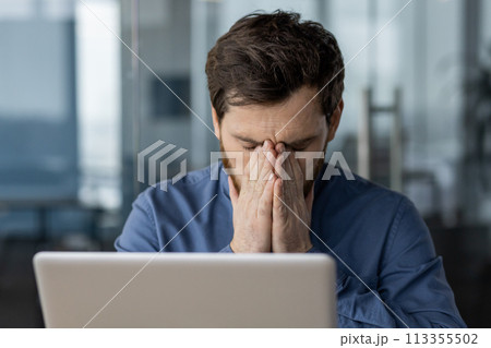 Overwhelmed male professional feeling stressed at work, sitting with hands covering his face in a modern office setting. 113355502