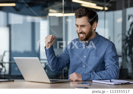 Young professional exults with a fist pump at his desk in a modern office setting, symbolizing success and achievement. 113355517