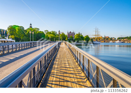 A vibrant day at Skeppsholmen Bridge in Stockholm, with clear skies and sunlight bathing the walkway, the calm water, and historic buildings in the distance. Stockholm, Sweden A vibrant day at Skeppsholmen Bridge in Stockholm, with clear skies and sunlight bathing the walkway, the calm water, and historic buildings in the distance. Stockholm, Sweden 113356262