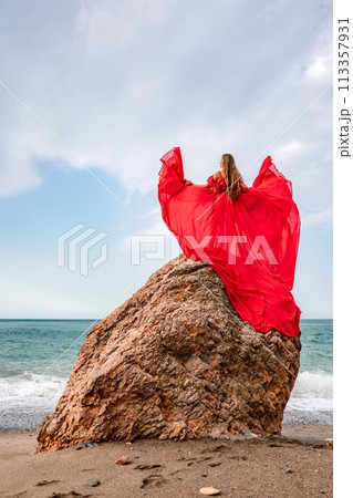 woman sea red dress. Woman with long hair on a sunny seashore in a red flowing dress, back view, silk fabric waving in the wind. Against the backdrop of the blue sky and mountains on the seashore. woman sea red dress. Woman with long hair on a sunny seashore in a red flowing dress, back view, silk fabric waving in the wind. Against the backdrop of the blue sky and mountains on the seashore. 113357931