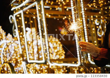 Woman holding sparkler night while celebrating Christmas outside. Dressed in a fur coat and a red headband. Blurred christmas decorations in the background. Selective focus 113358430