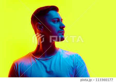 Serious young athletic man posing in casual attire in neon light against vibrant yellow studio background. Serious young athletic man posing in casual attire in neon light against vibrant yellow studio background. 113360177