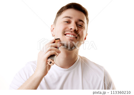 Young man in white T-shirt shaving his beard with electric razor against white studio background. Hygiene. 113360210