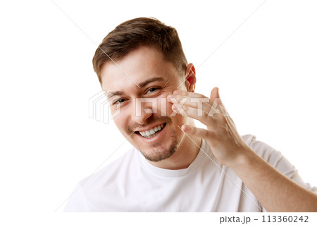 Young man in T-shirt applying cosmetic organic facial-care product smiling looking at camera against white background. 113360242