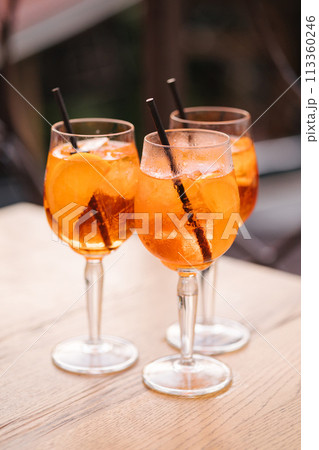 Three glasses with orange alcoholic drink on wooden table. Black straw in a drink. Aperol spritz cocktail in a wine glass Three glasses with orange alcoholic drink on wooden table. Black straw in a drink. Aperol spritz cocktail in a wine glass 113360246