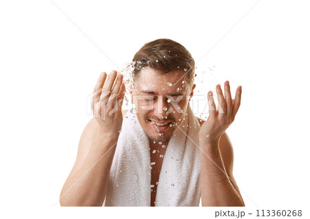 Young man with towel on shoulders washing his face with splashes of water in motion against white studio background. 113360268