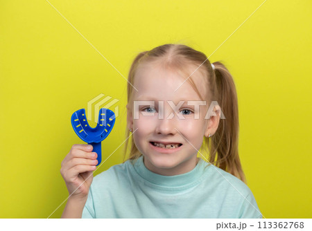Smiling girl aged seven years old with a blue dental impression tray on a yellow background. The concept of making impressions of the dental jaw in children in orthodontics. Making veneers and Smiling girl aged seven years old with a blue dental impression tray on a yellow background. The concept of making impressions of the dental jaw in children in orthodontics. Making veneers and 113362768