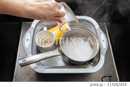 A woman's hand pours hot water into a basin with dirty dishes and environmentally friendly oxygen bleach. Washing dishes from grease and dirt. A woman's hand pours hot water into a basin with dirty dishes and environmentally friendly oxygen bleach. Washing dishes from grease and dirt. 113362854