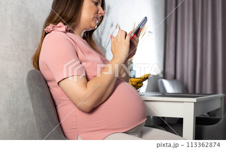 Pregnant girl with a mobile phone in the kitchen. The concept of the negative impact of phone radiation on the fetus in the womb. Pregnant girl with a mobile phone in the kitchen. The concept of the negative impact of phone radiation on the fetus in the womb. 113362874