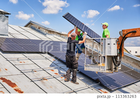 Workers lifting up photovoltaic solar panel on metal rooftop of house with assistance of crane lift. Men installers installing photovoltaic solar modules outdoors. Renewable energy generation concept. Workers lifting up photovoltaic solar panel on metal rooftop of house with assistance of crane lift. Men installers installing photovoltaic solar modules outdoors. Renewable energy generation concept. 113363263