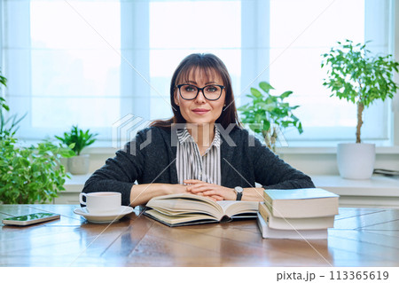 Middle-aged woman reading books, sitting at home at table, smiling looking at camera 113365619