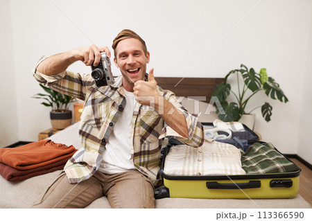 Portrait of handsome tourist, male model holding a camera, sitting near opened suitcase with clothes, man going on vacation, packing for summer holiday retreat Portrait of handsome tourist, male model holding a camera, sitting near opened suitcase with clothes, man going on vacation, packing for summer holiday retreat 113366590